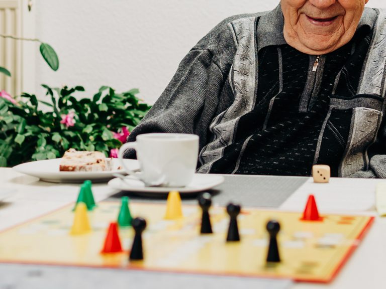 In der solitas Tagespflege sind Gesellschaftsspiele sehr beliebt. Auf dem Bild ist ein Tisch mit einem Mensch-ärgere-dich-nicht-Spiel, im Hintergrund ein lachender Senior im grau-schwarzen Pullover.