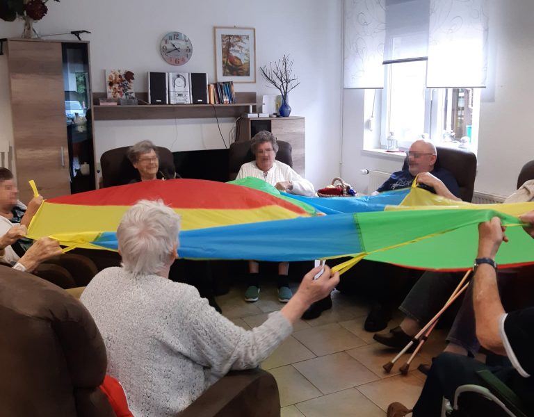 solitas Schwungtuchübungen: Mehrere Senioren sitzen im Kreis und bewegen mit den Armen ein buntes Schwungtuch hoch und runter.